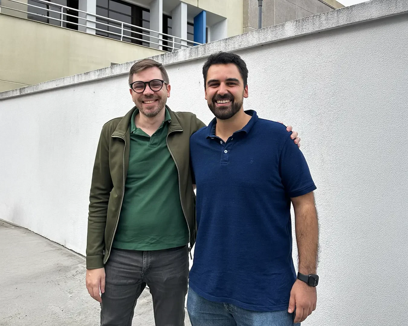 Bruno and Guilherme standing outdoors in front of a white wall, smiling at the camera. The man on the left is wearing glasses, a green polo shirt, and a green jacket. The man on the right is wearing a navy blue polo shirt and has his arm around the other man.