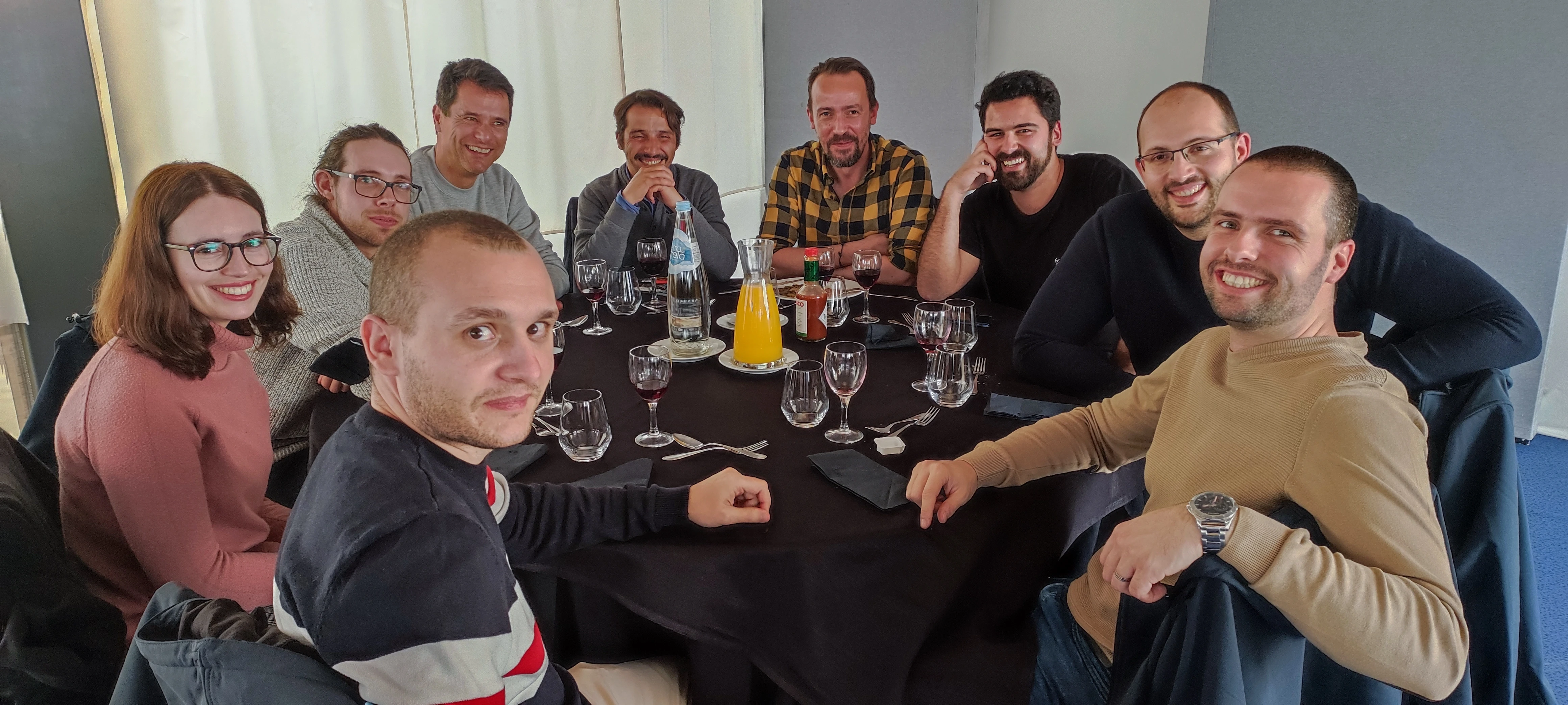 A group of nine people sitting around a round table set for a meal, smiling at the camera. The table has glasses, cutlery, a carafe of orange juice, and bottled water. The group appears relaxed and happy in a bright indoor setting.