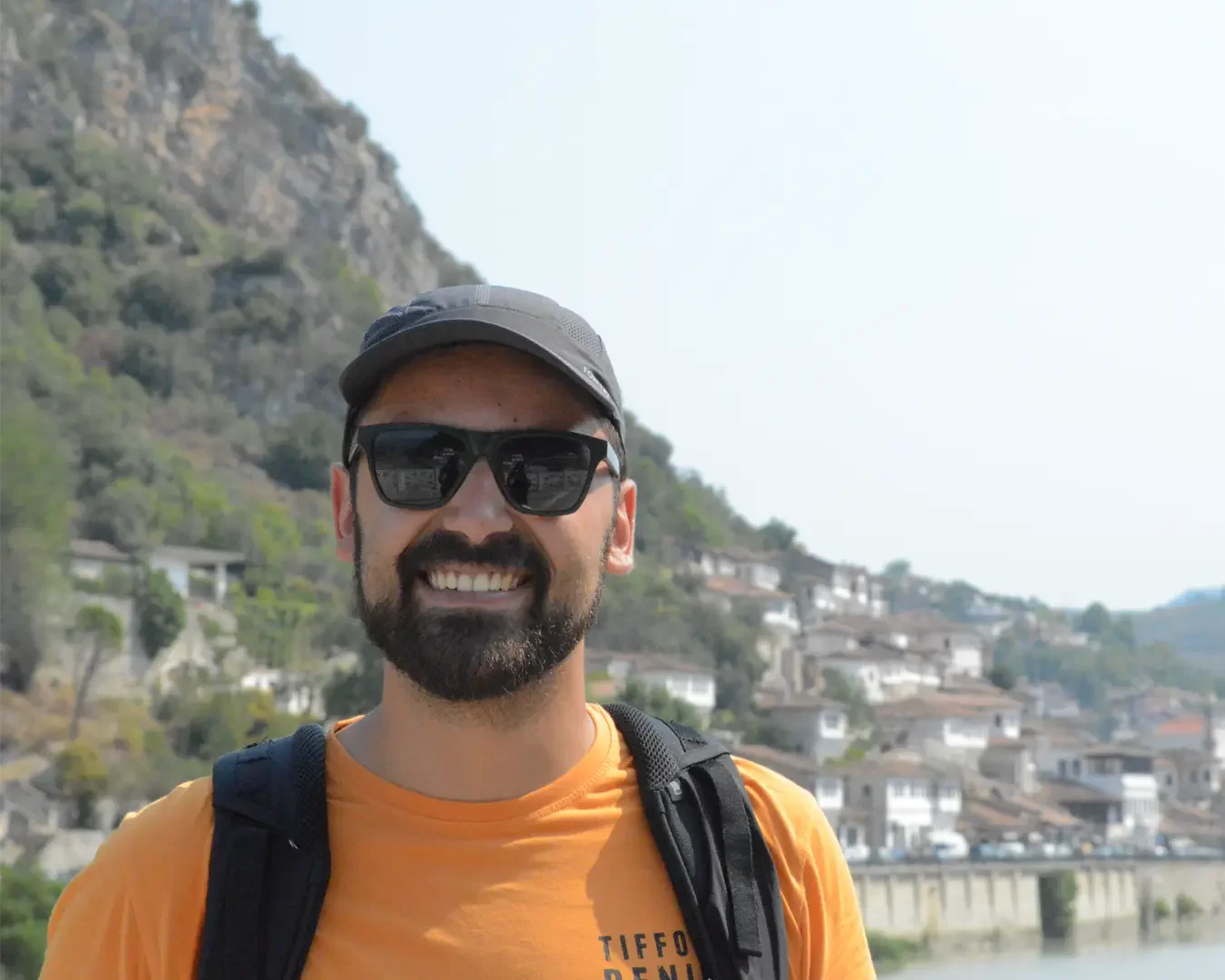 A smiling man wearing sunglasses, a black cap, and an orange T-shirt stands outdoors with a hillside village and river behind him.