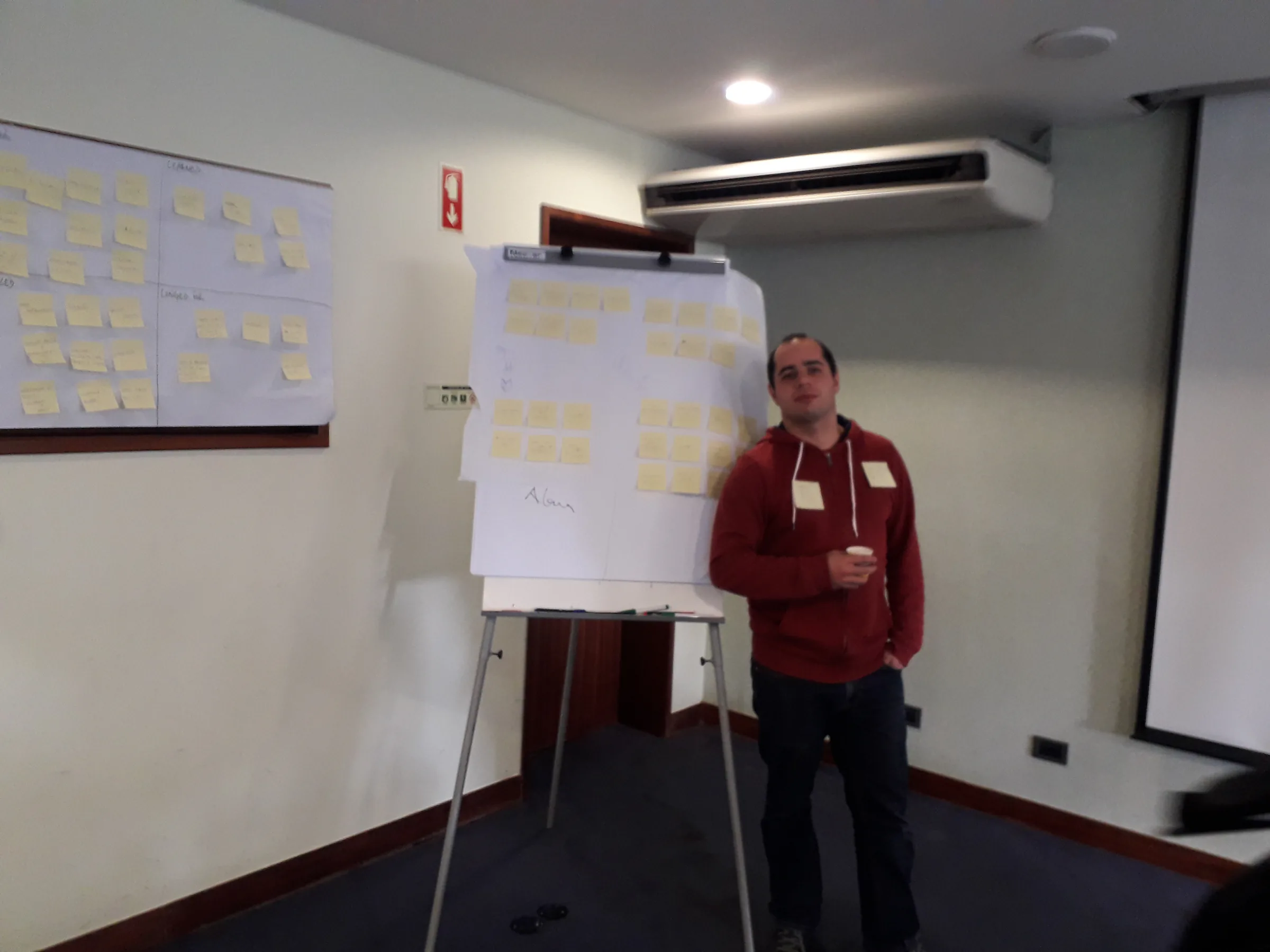 João Franco, Focus BC Alumni in a red hoodie standing next to a flipchart covered in yellow sticky notes, presenting or explaining something during a workshop. More sticky notes are visible on the wall-mounted paper behind him.