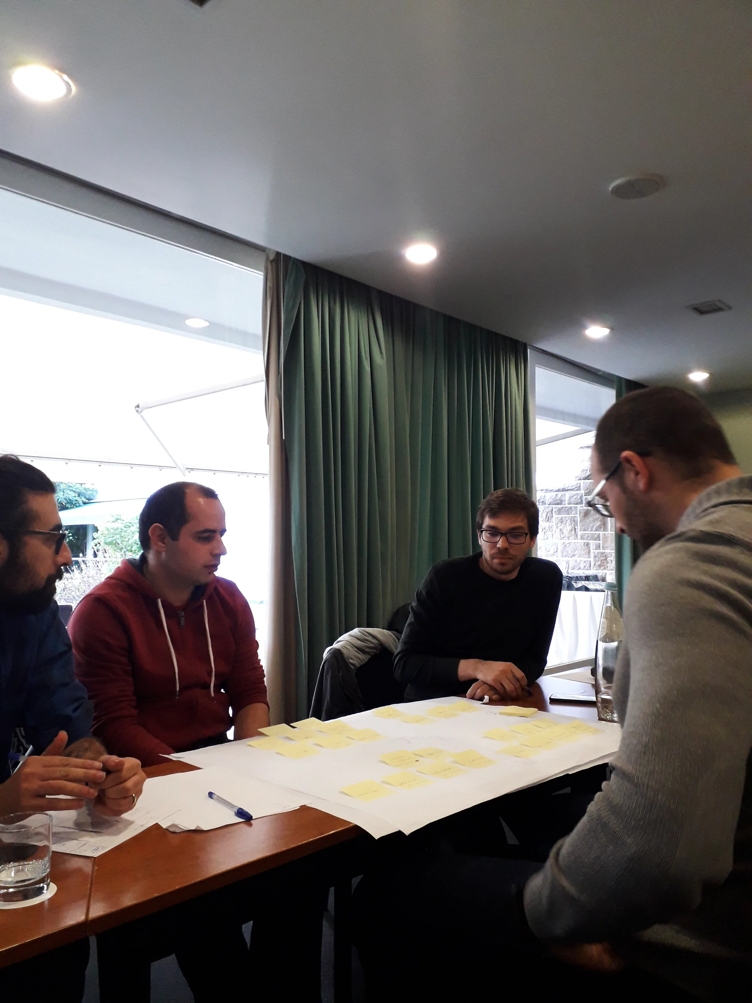 João Franco, Focus BC Alumni and other Focus BC team members sitting around a table in a meeting room, discussing and working with sticky notes on a large sheet of paper. One of them holds a pen while others look attentively at the notes.