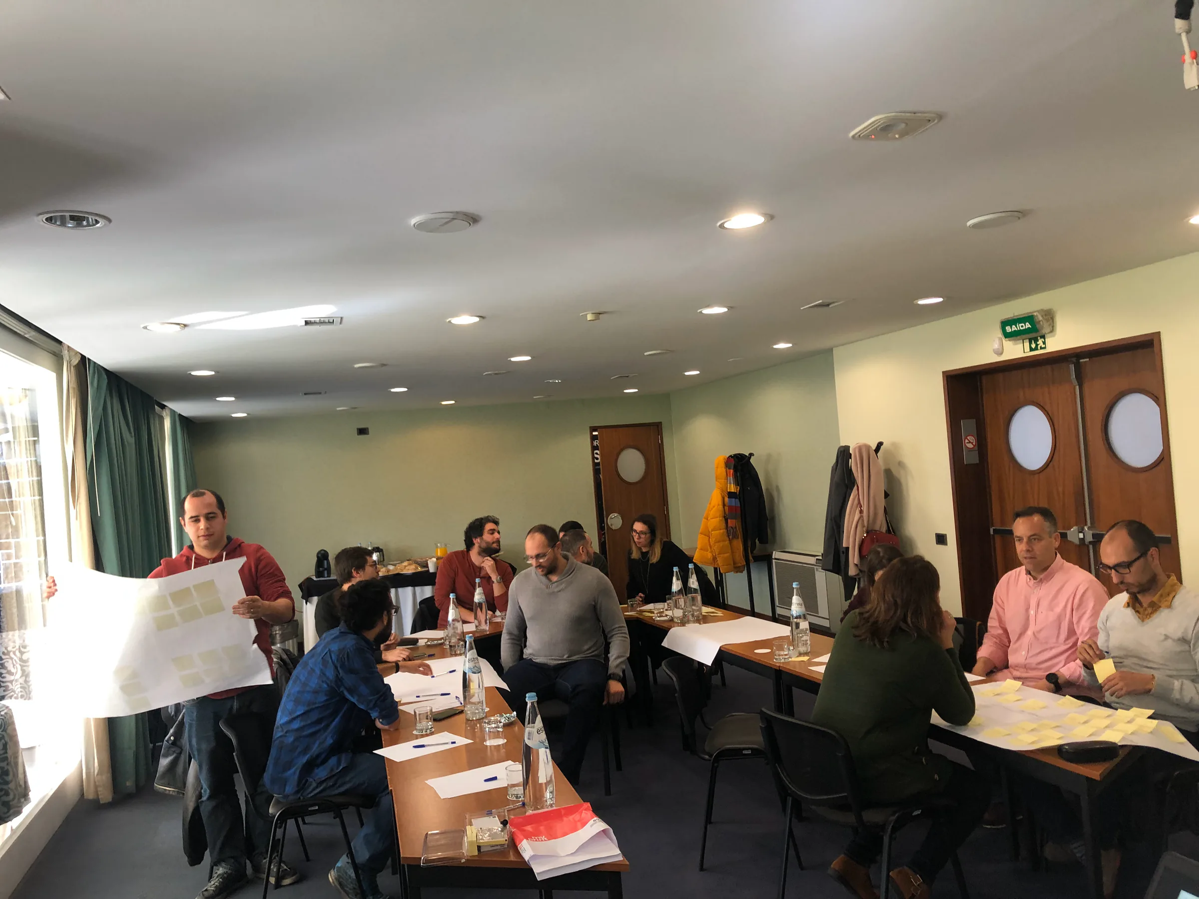 Focus BC team participating in a  team activity in a conference room. Several tables are filled with groups working with paper and sticky notes. One man stands at the front holding a large sheet of paper covered in notes.