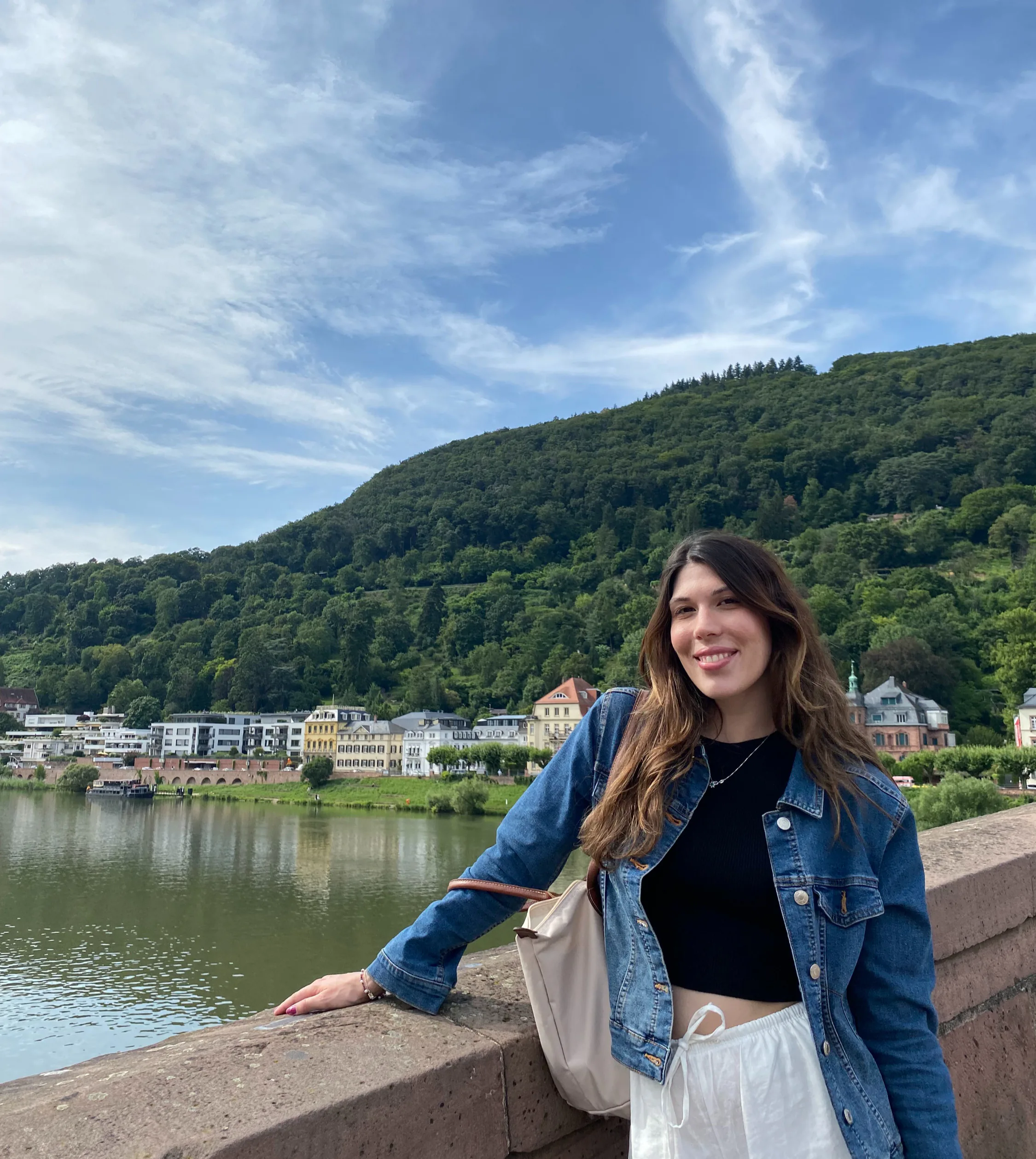 Rita standing on a stone bridge by a calm river, smiling at the camera. She is wearing a denim jacket over a black top and white trousers, with a light-colored shoulder bag. In the background, there are hillside forests and a row of buildings along the riverbank under a blue sky with light clouds.