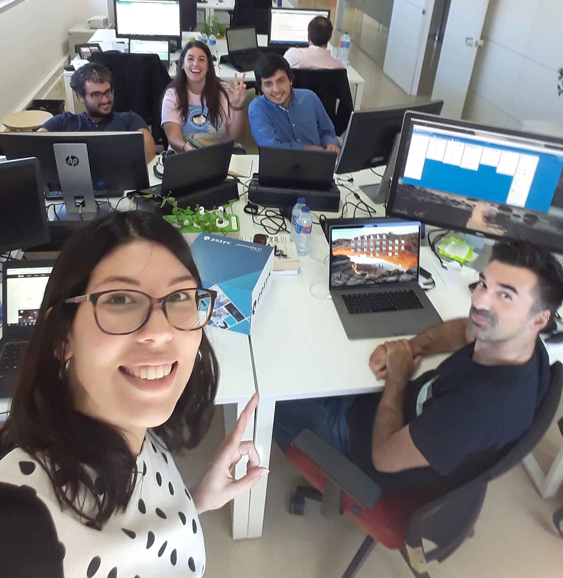 Selfie taken in an office with a group of colleagues working at desks with multiple monitors and laptops. In the foreground, a woman wearing glasses smiles at the camera and points toward the team. Around the table, several coworkers sit and look up, one making a peace sign. The desks are covered with computers, cables, notebooks, and a water bottle, creating a busy work environment.