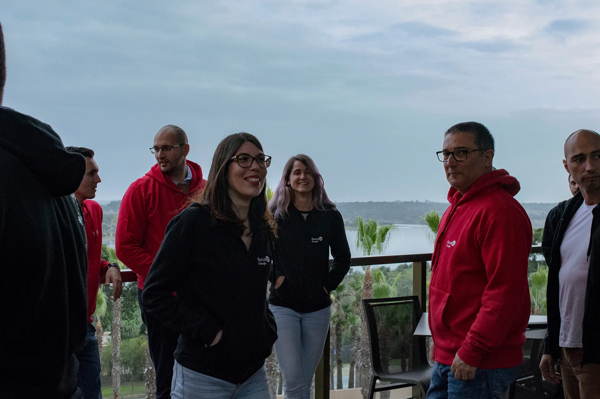 Group of colleagues standing on an outdoor terrace overlooking a river and palm trees. Some are wearing red hoodies with a company logo, while others wear black jackets. They are casually gathered and chatting, with a scenic landscape and cloudy sky in the background.