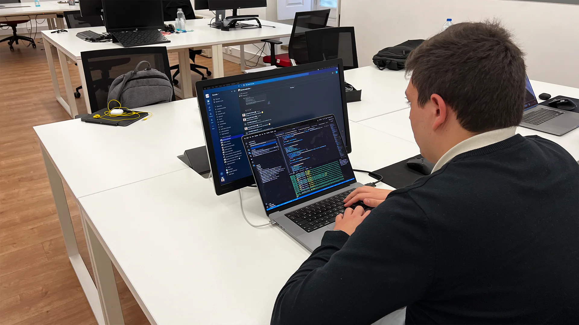 Software developer working on a laptop connected to an external monitor in a modern open-plan office.