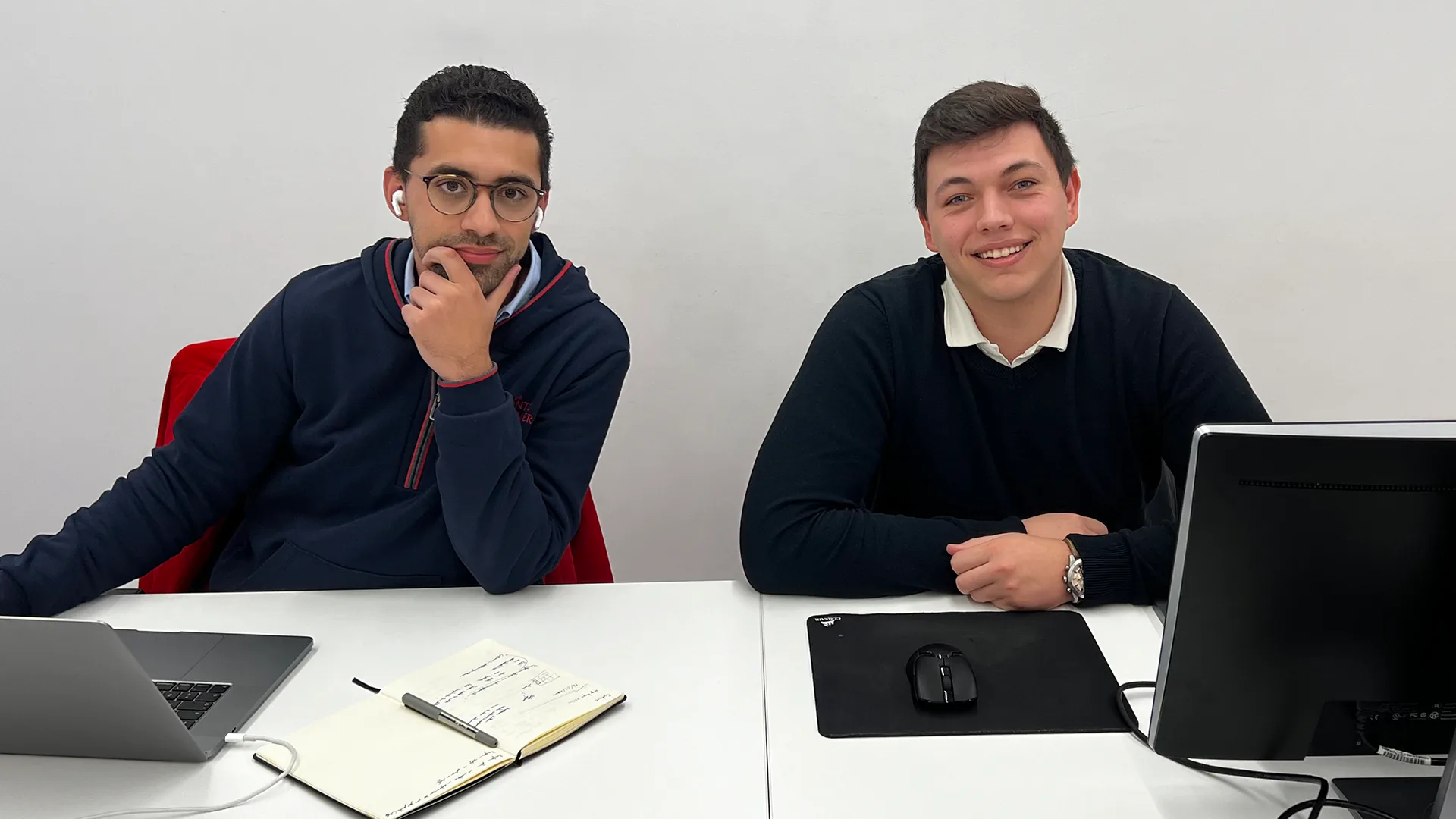 Two colleagues seated at a desk in an office, smiling at the camera with laptops and work materials in front of them.