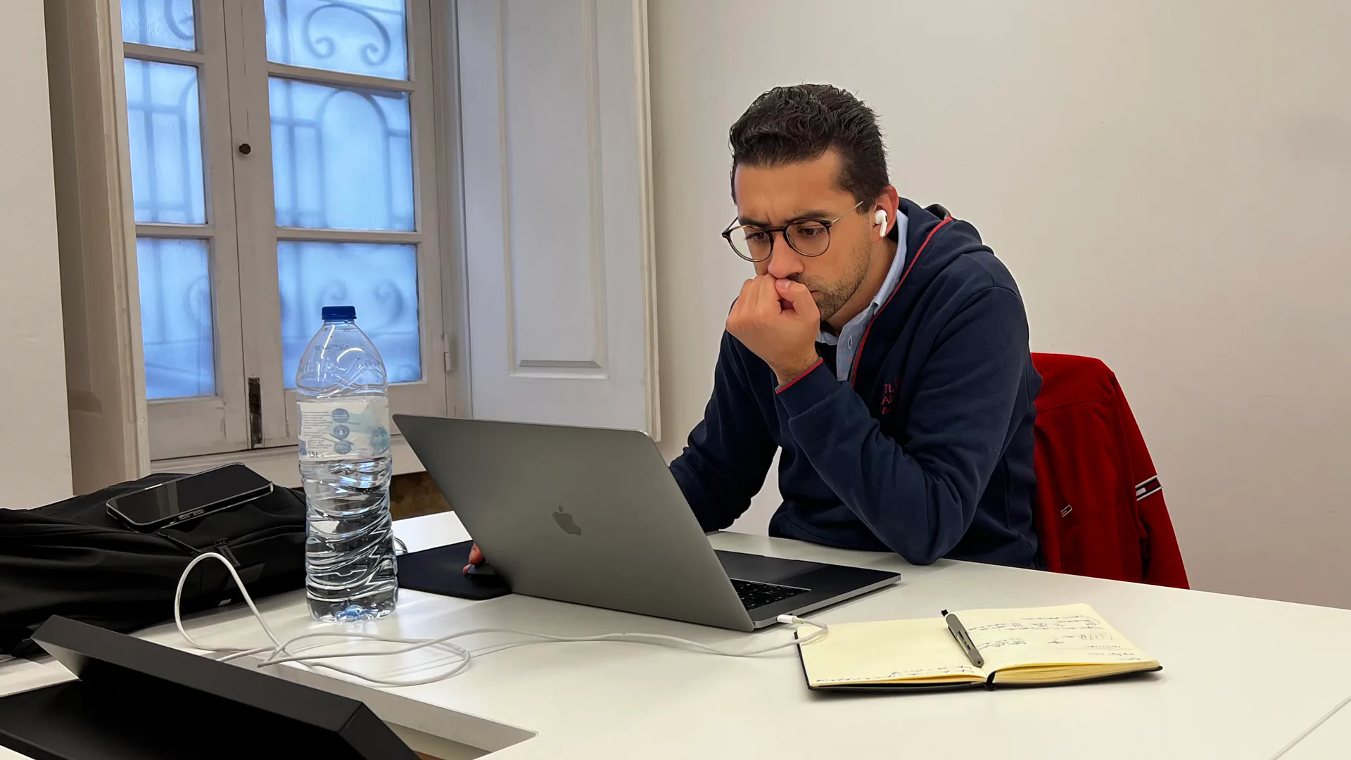 Focused professional working on a laptop at a desk, with a notebook and water bottle beside him in a bright office space.