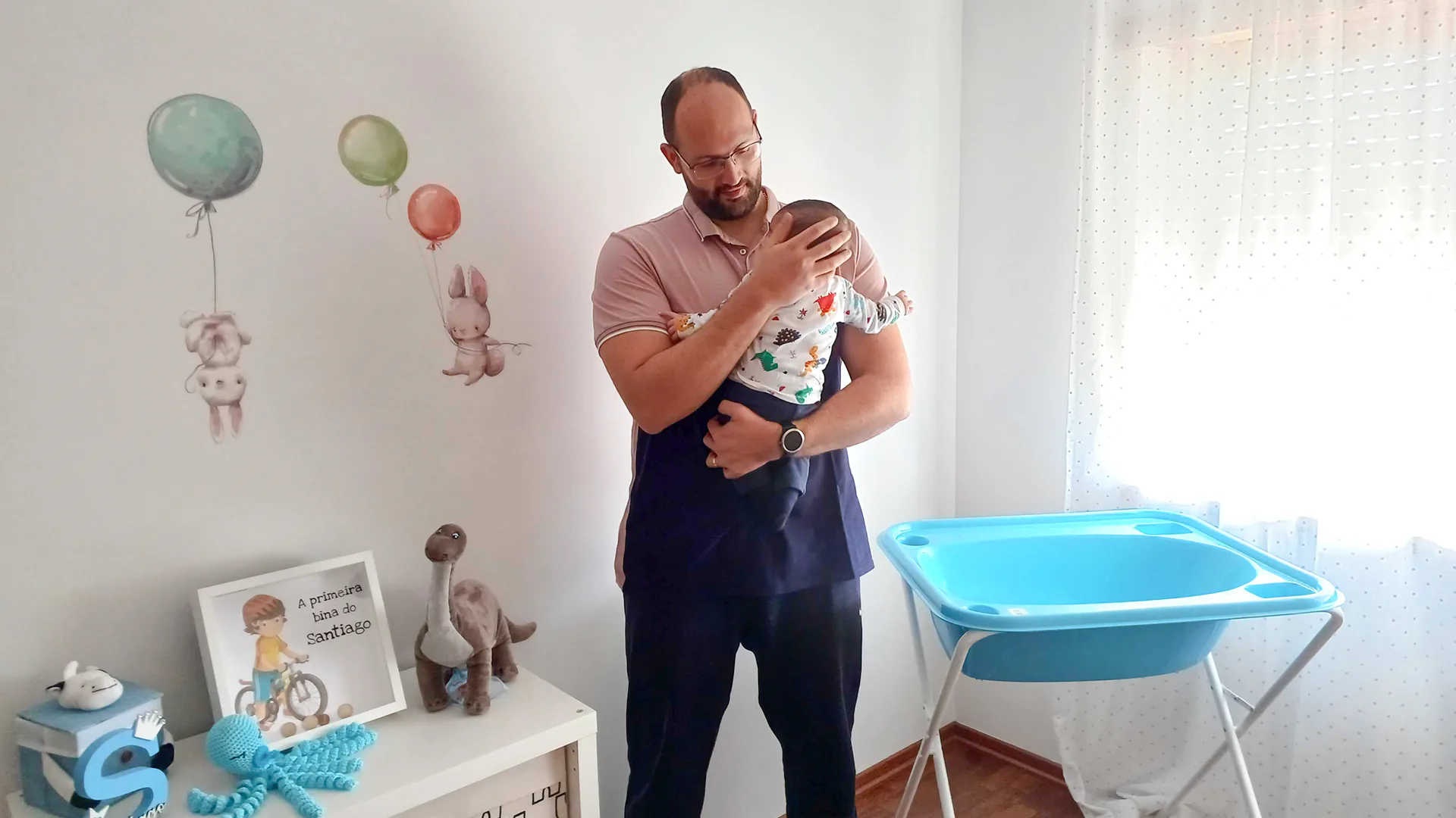 Father gently holding and comforting his baby in a bright nursery decorated with soft toys, balloons, and baby furniture.