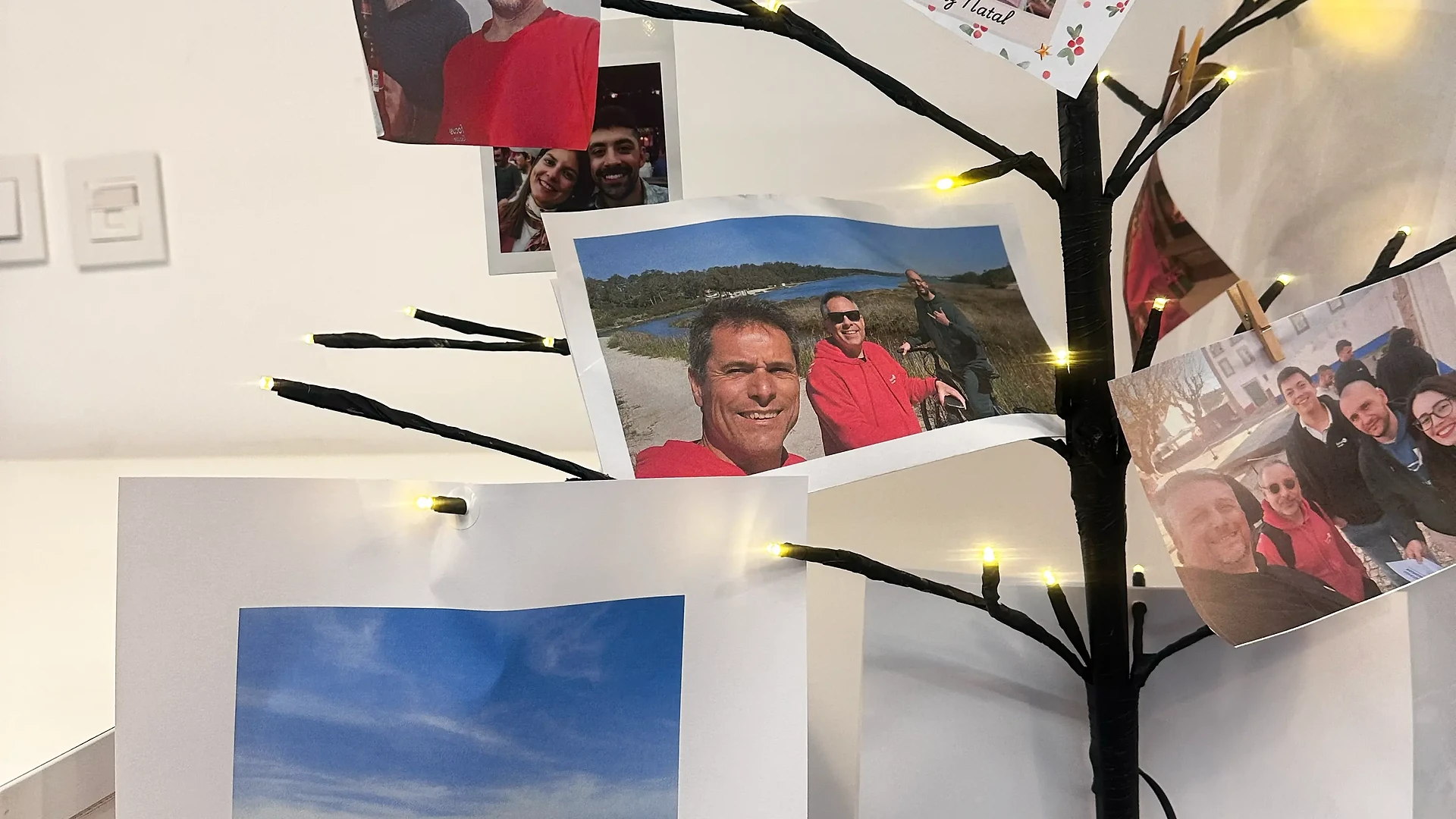 A small decorative Christmas tree with warm white lights, displaying printed team photos and holiday messages clipped to its branches.