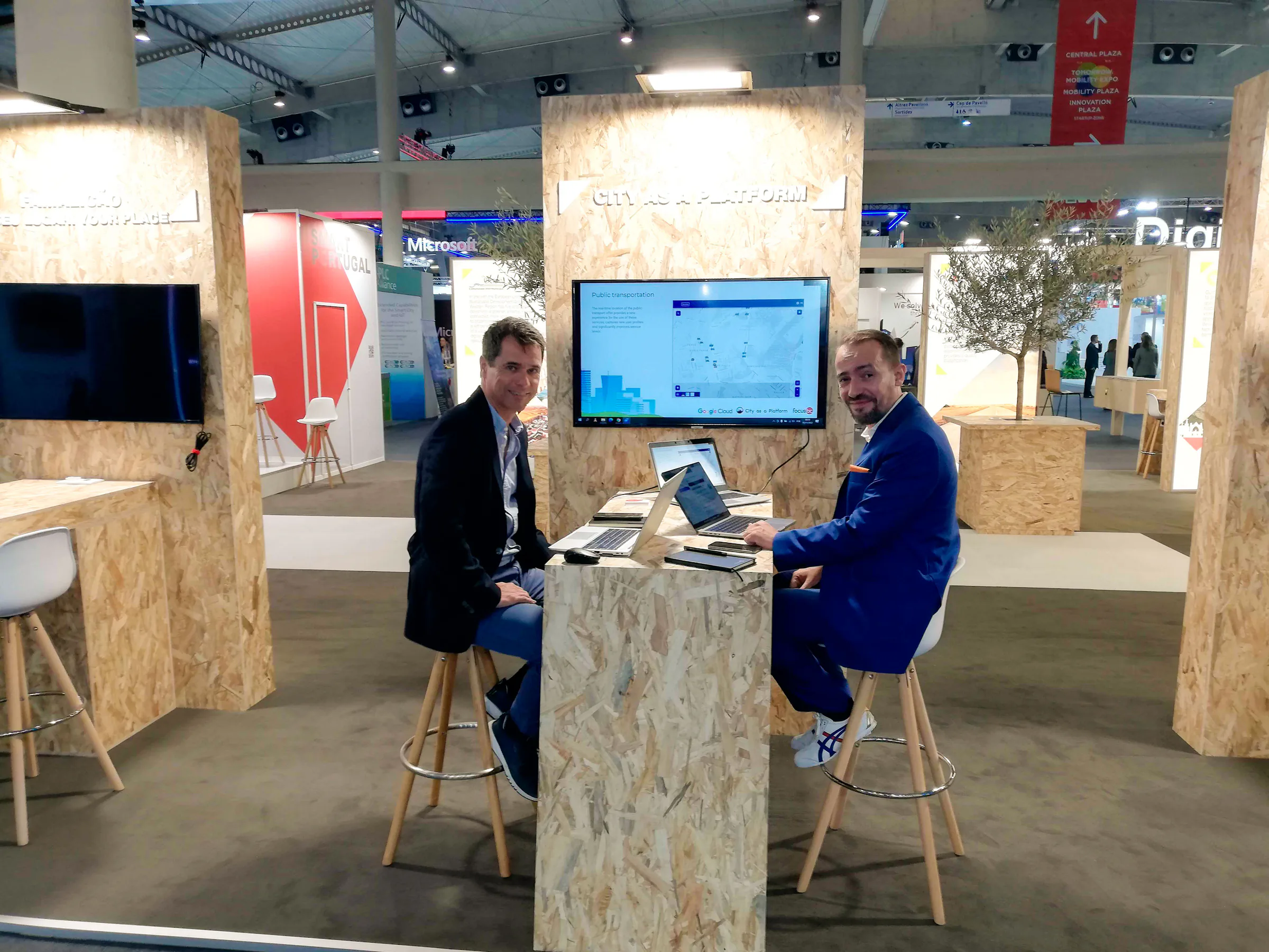 Vasco and Sandro seated at a trade show booth demonstrating the City as a Platform solution on laptops, with a screen displaying a smart city interface behind them.