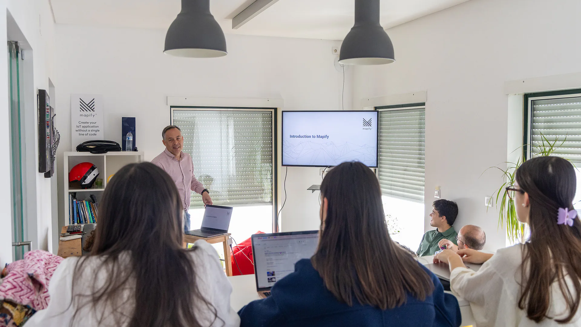 Mario Sobral stands in front of a group, gesturing with both hands as he leads a session. A poster behind him reads “Mapify – Create your IoT application without a single line of code.” The room is bright, with a laptop and books on a shelf.