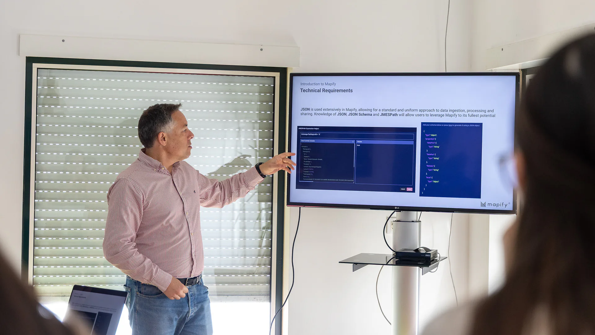 A cozy and bright room set up for a workshop with colorful beanbags arranged in a semicircle facing a TV screen displaying a presentation slide titled “Introduction to Mapify.” A laptop showing the same slide rests on a wooden stool. Sunlight filters through partially closed blinds.