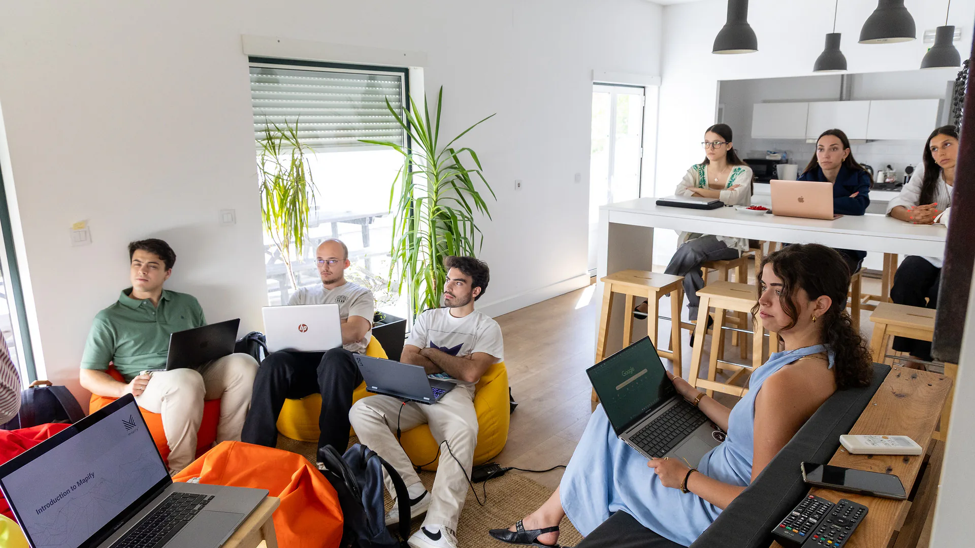 A cozy and bright room set up for a workshop with colorful beanbags arranged in a semicircle facing a TV screen displaying a presentation slide titled “Introduction to Mapify.” A laptop showing the same slide rests on a wooden stool. Sunlight filters through partially closed blinds.