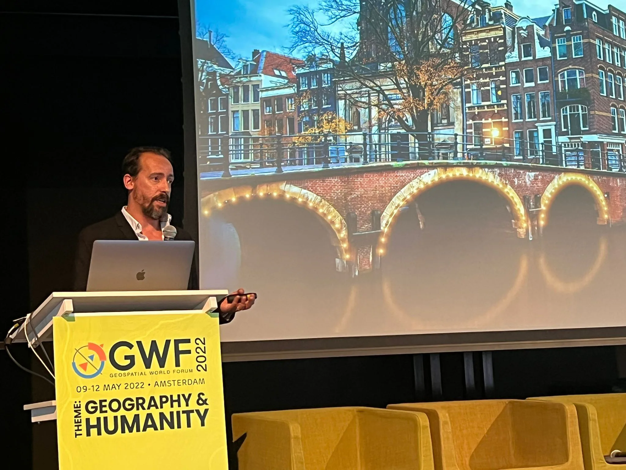 Speaker presenting at the Geospatial World Forum 2022 in Amsterdam, standing at a podium labeled “GWF 2022 – Geography & Humanity,” with an image of Amsterdam’s canals projected behind him.