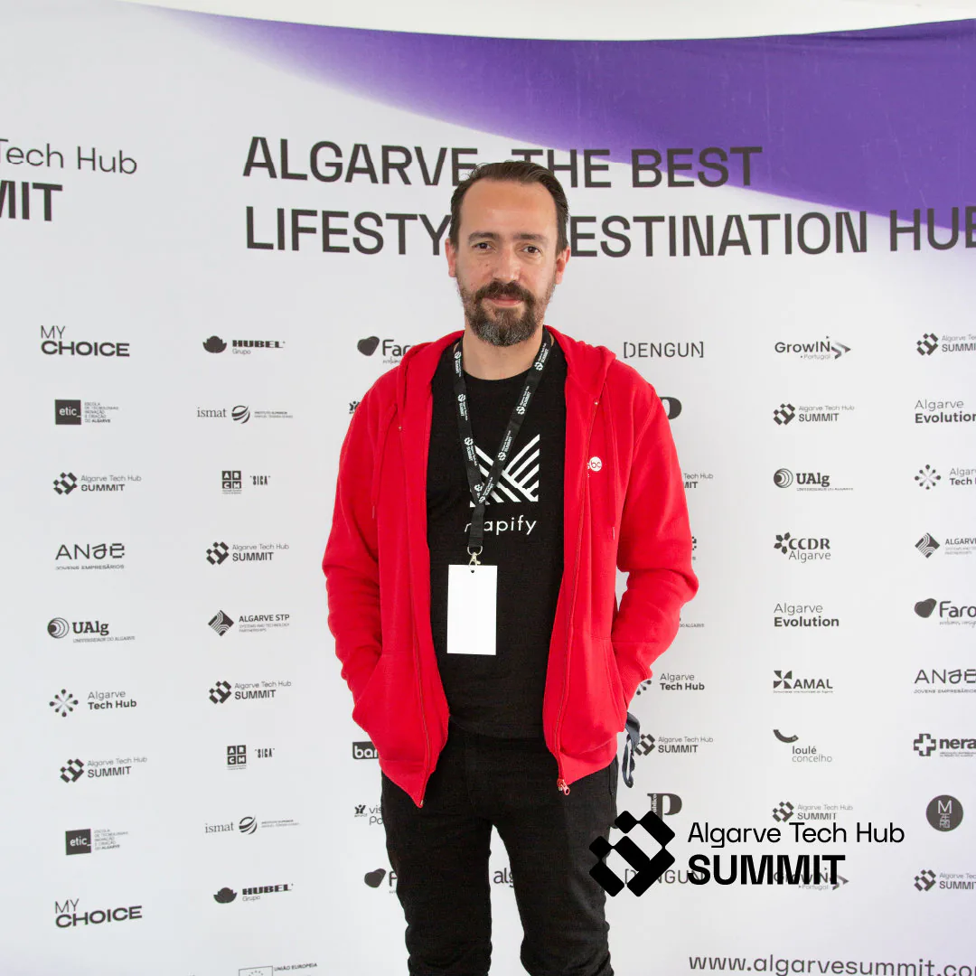 Event attendee wearing a red hoodie and Mapify T-shirt posing in front of an Algarve Tech Hub Summit backdrop with sponsor logos.
