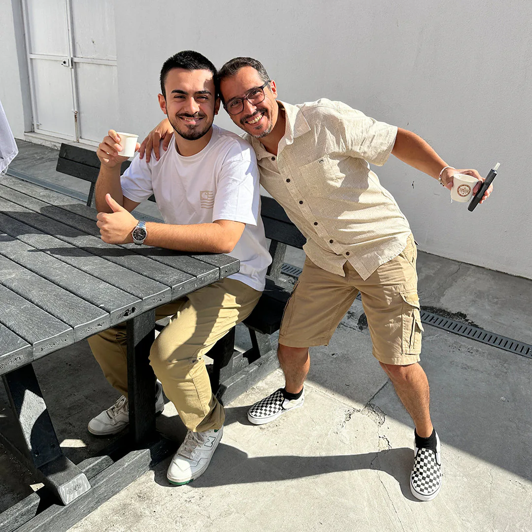 Two men smiling outdoors beside a wooden picnic table, holding small coffee cups. One sits wearing a white t-shirt and beige pants, while the other stands leaning in with an arm around his shoulder, wearing a light short-sleeve shirt and beige shorts.