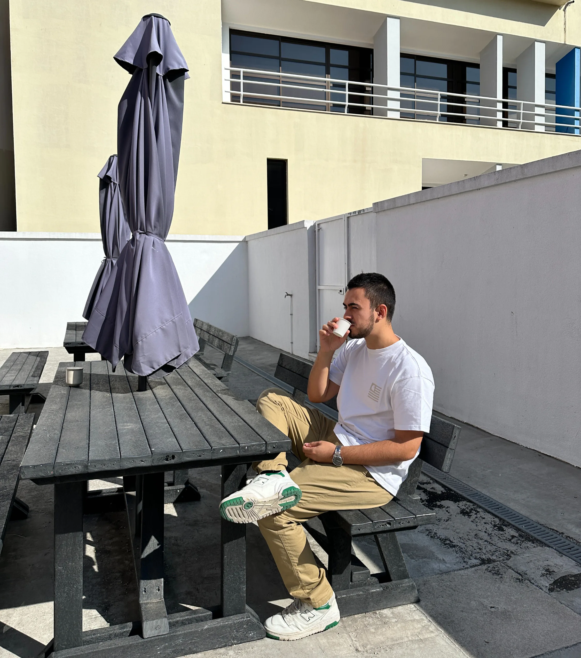 Tomás sitting alone at an outdoor wooden picnic table, drinking a small coffee cup in bright sunlight, with a closed patio umbrella and modern building facade in the background.