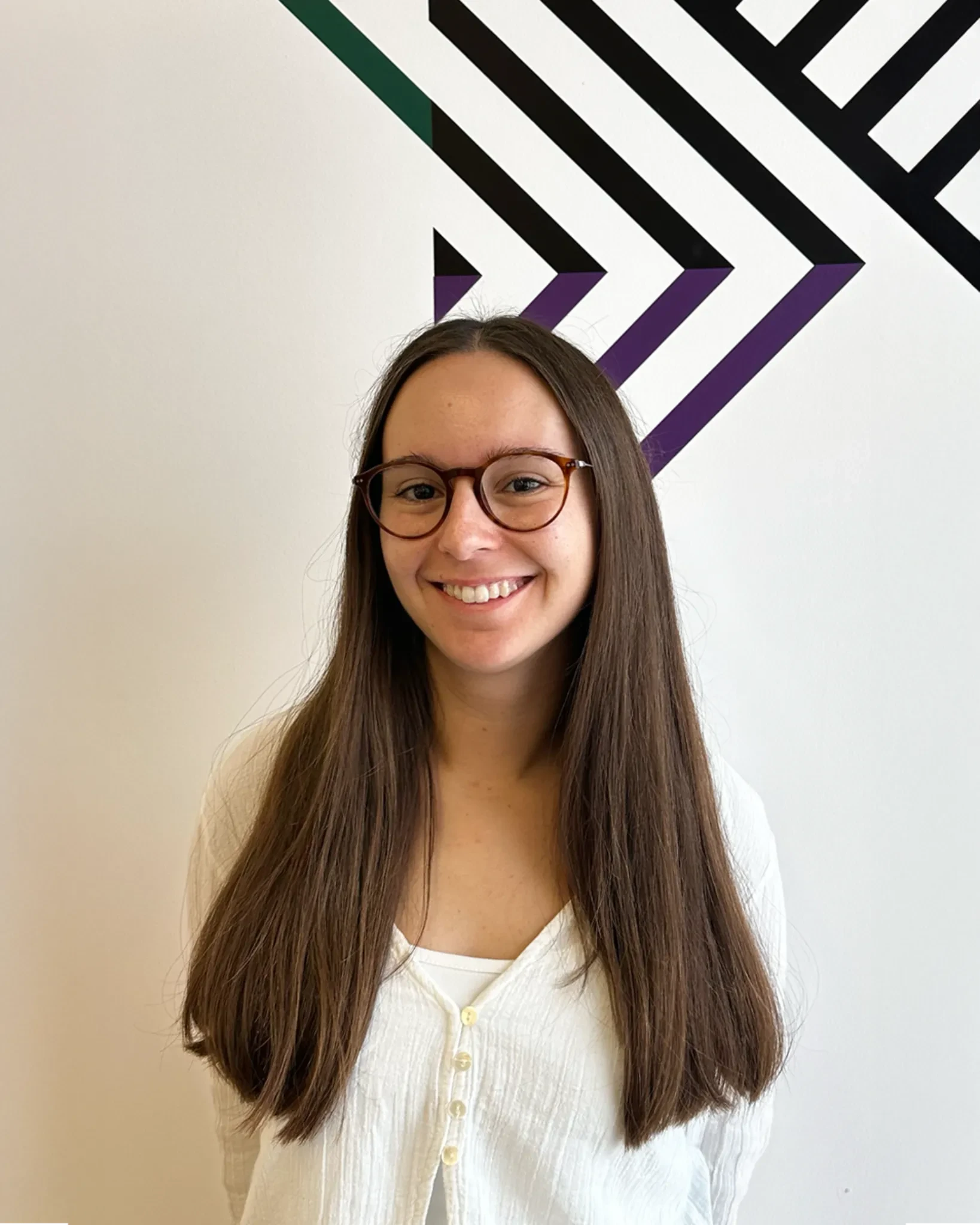 Smiling woman with long brown hair and round glasses, wearing a white top, standing in front of a white wall with a bold black and purple geometric pattern.