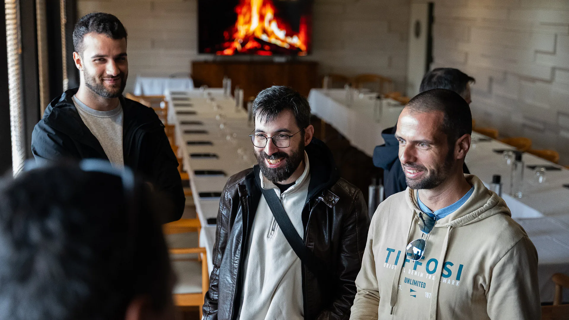 Man wearing a red hoodie and sunglasses standing with arms crossed outdoors, with a walkway, parked cars, and people in the background.