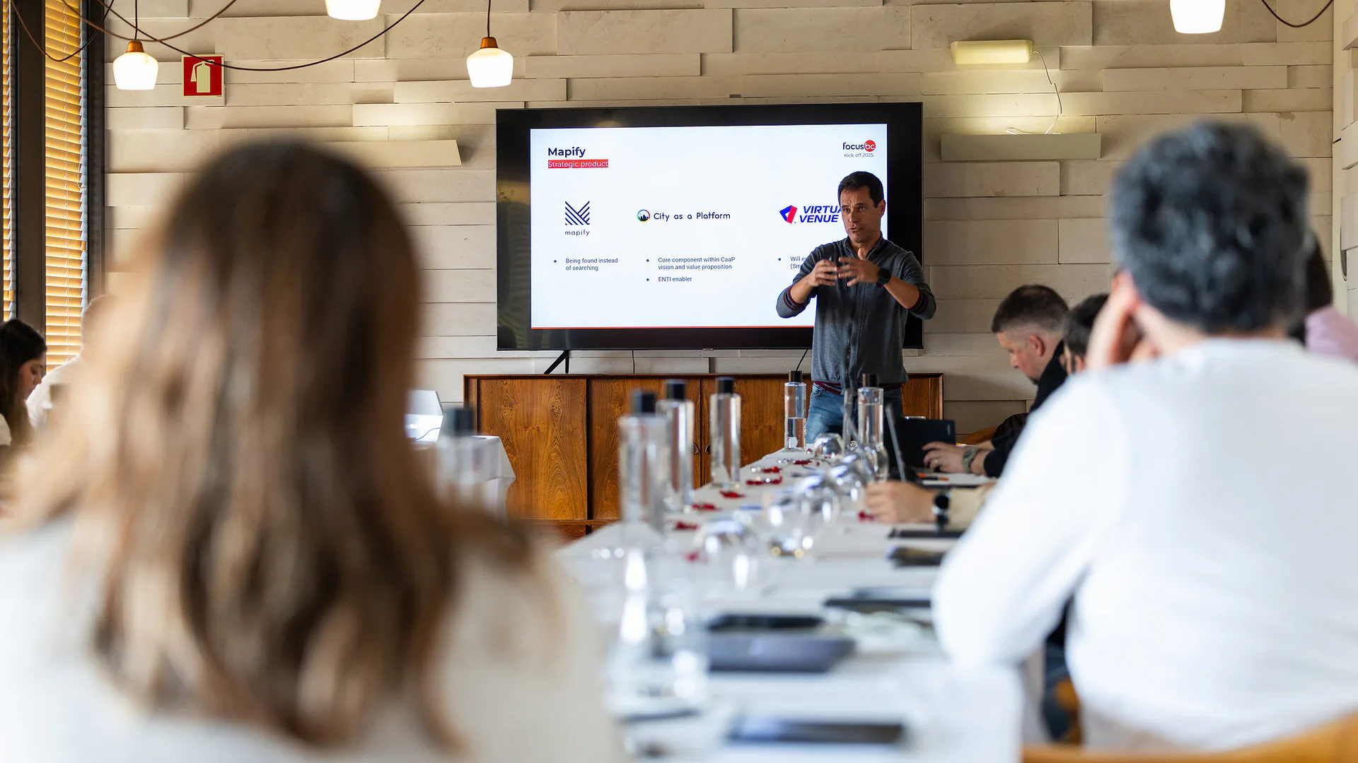 Three men smiling and talking indoors in a dining room set with long tables and chairs, with a large screen displaying a fireplace in the background.