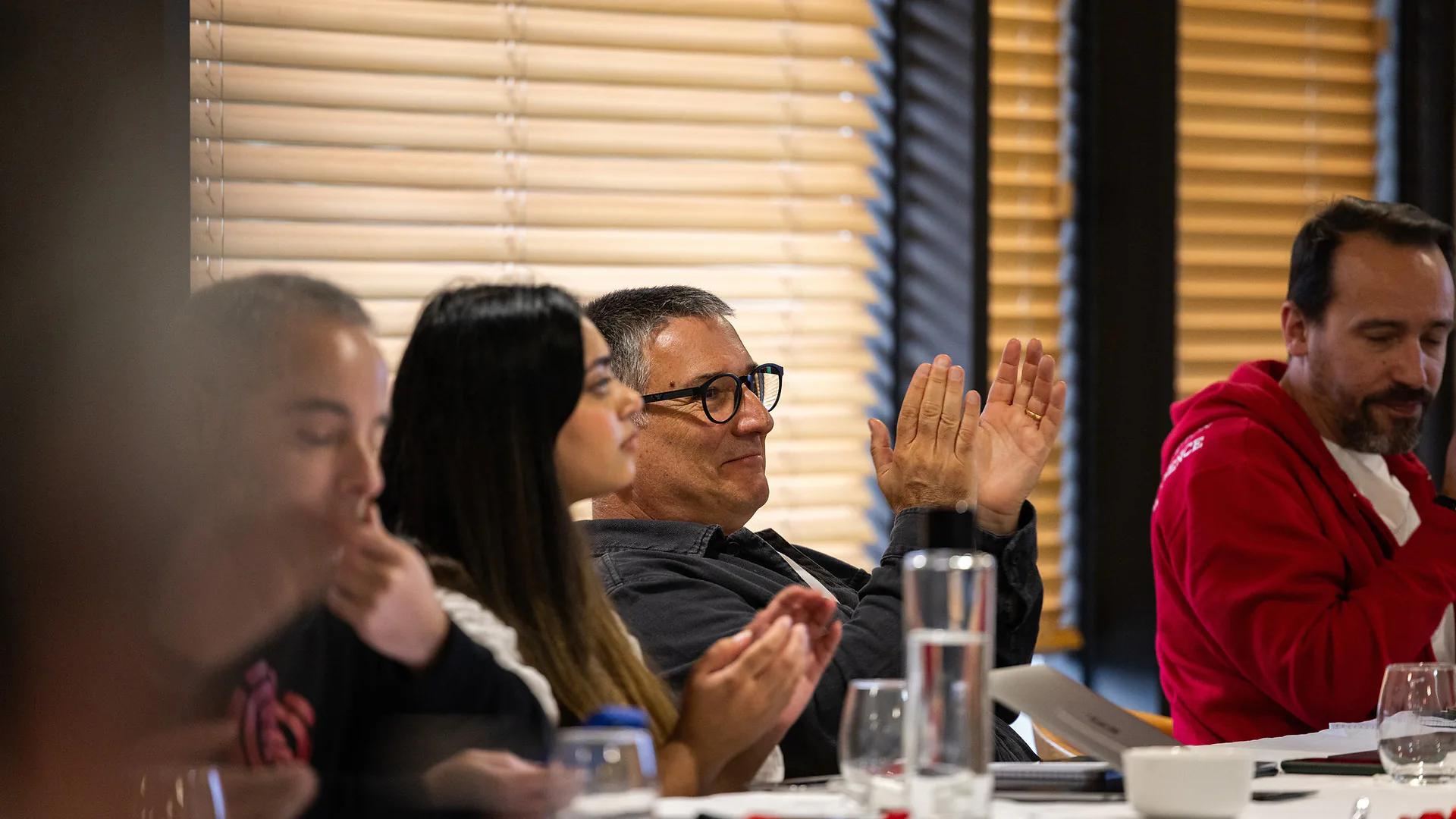 Wide view of a meeting room with people seated around long tables while a presenter speaks at the front beside a screen.
