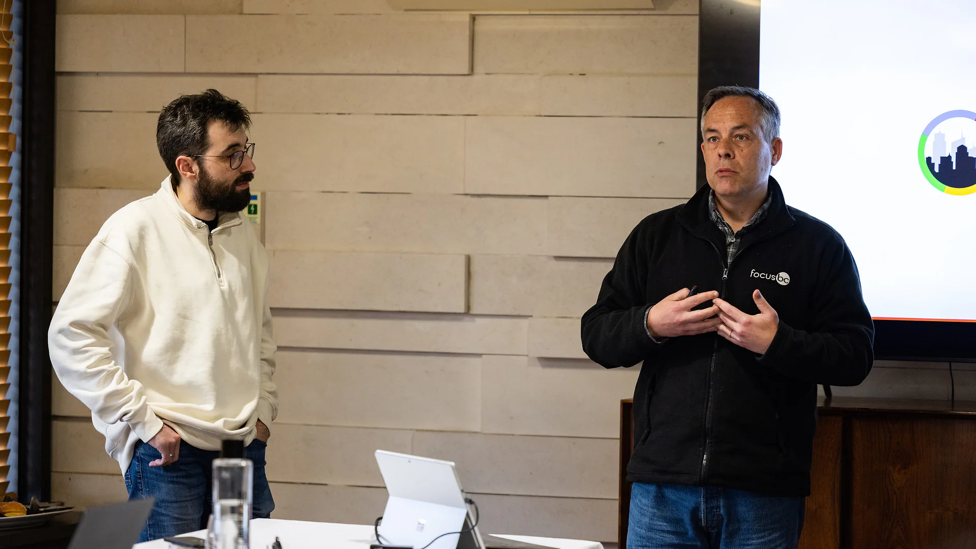 Wide view of a meeting room with people seated around long tables while a presenter speaks at the front beside a screen.