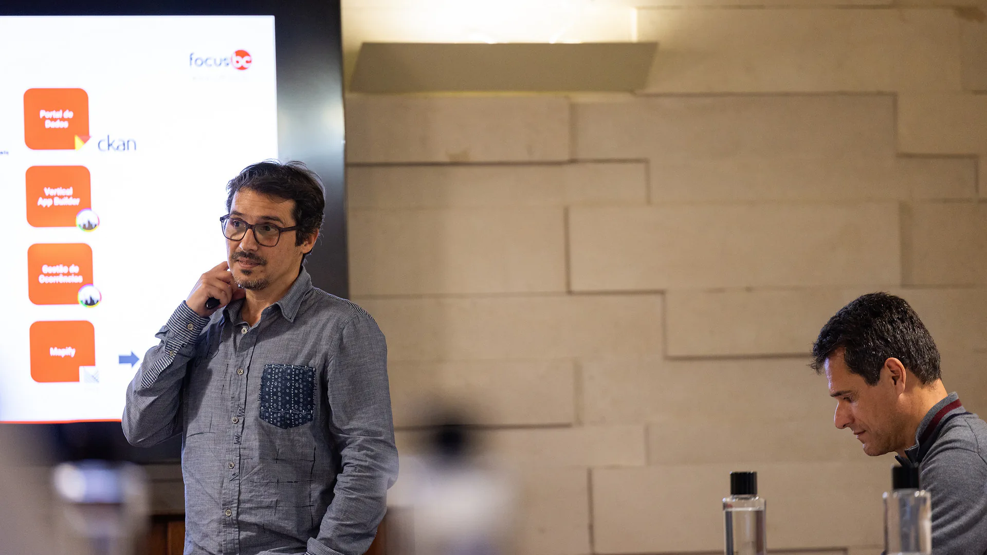 Presenter standing in front of a screen displaying slides, speaking to an audience seated at long tables.