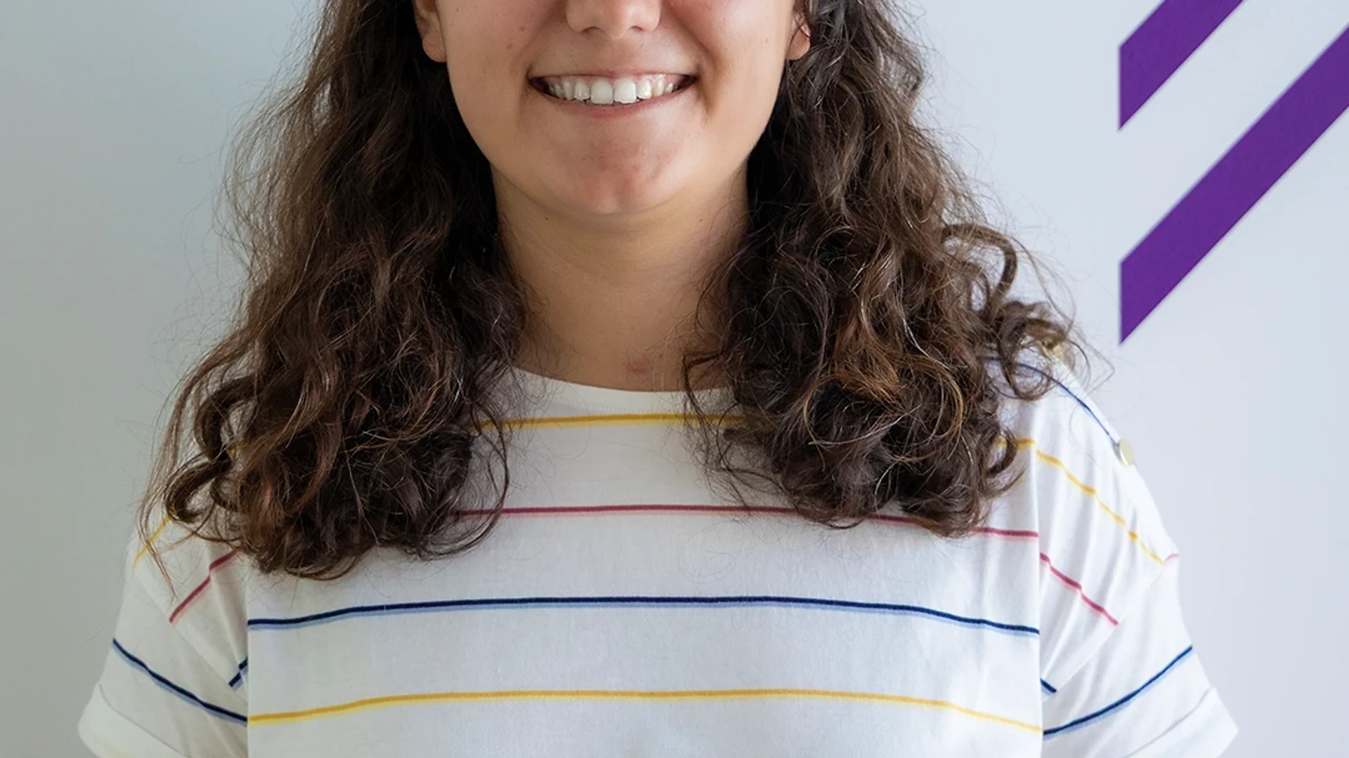 Joana sitting on a green bean bag in the office lounge, smiling toward the camera with colorful bean bags and a TV in the background.