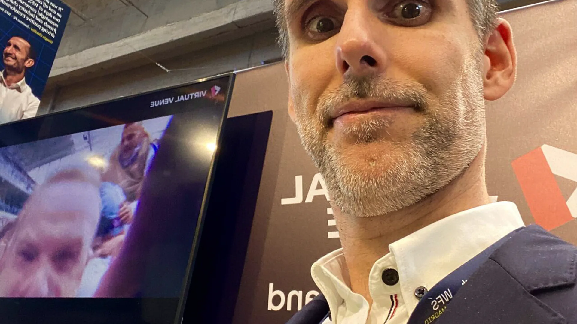 A man sitting at his workstation in a modern office, working on a laptop connected to multiple screens. On the wall behind him, the text reads “Hello world, we are mapify.” Another person is seated across the table, engaging in conversation.
