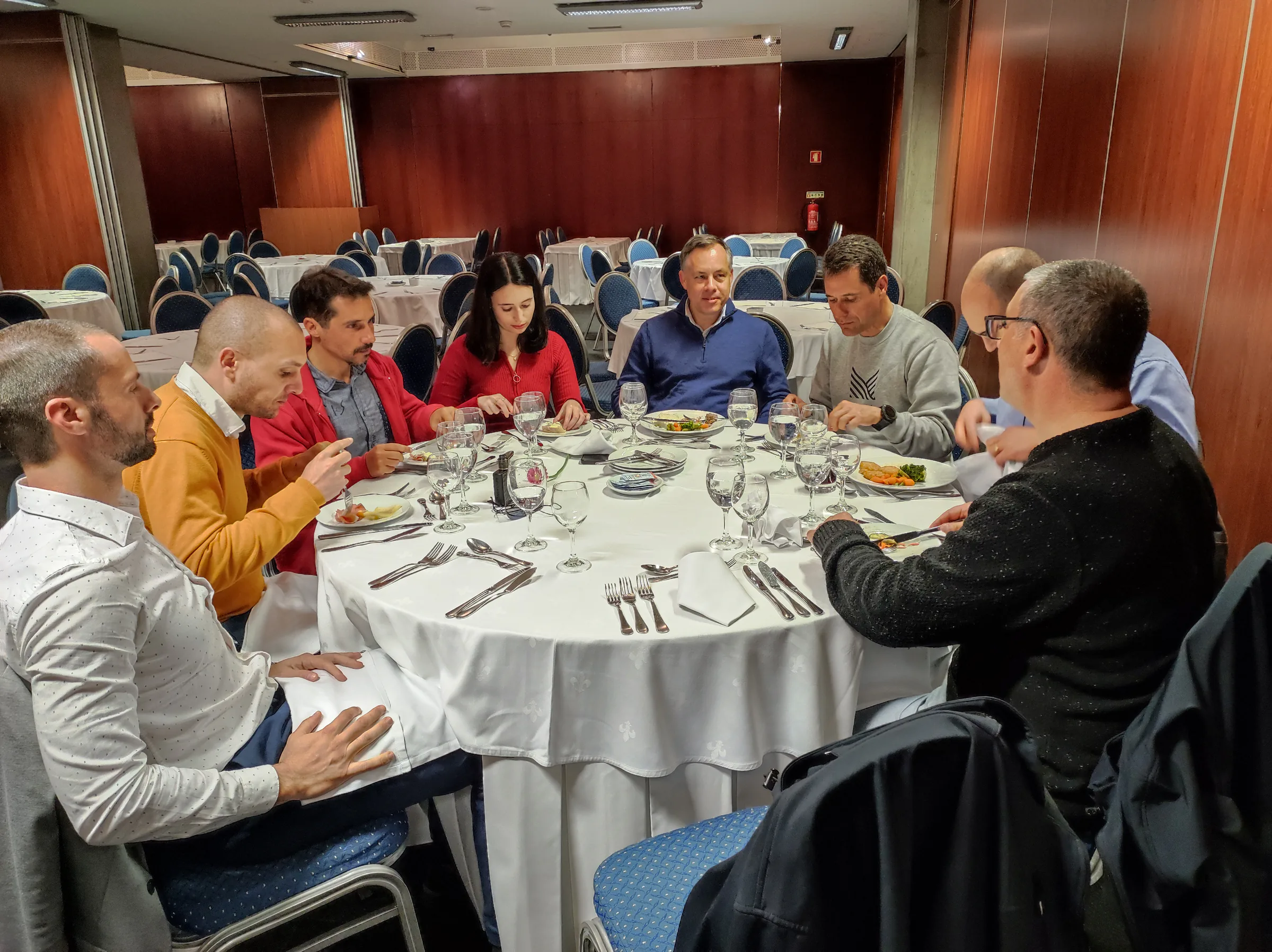 Man in a grey sweater presenting in a meeting room, standing beside a projector and addressing colleagues seated at tables.