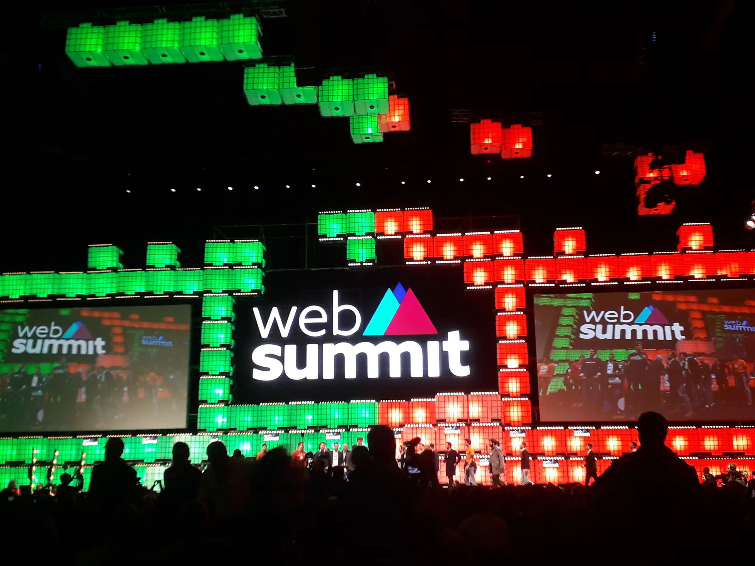Web Summit main stage with large illuminated “web summit” signage in red and green lights, speakers on stage and audience silhouettes in the foreground.