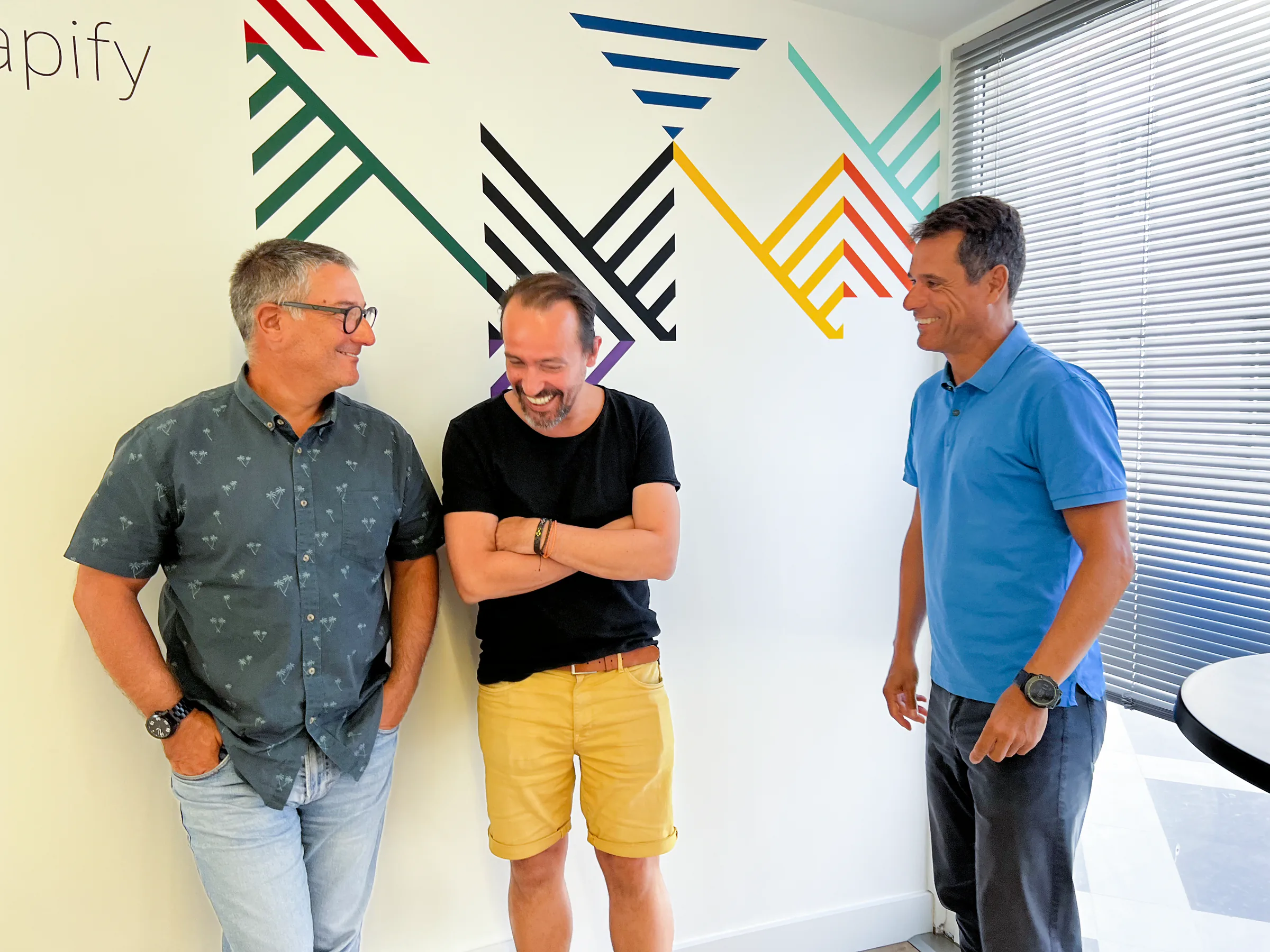 Founders standing indoors in front of a white wall with colorful geometric line graphics, smiling and chatting casually; one wears a patterned short-sleeve shirt and glasses, another a black t-shirt with yellow shorts, and the third a blue polo shirt.