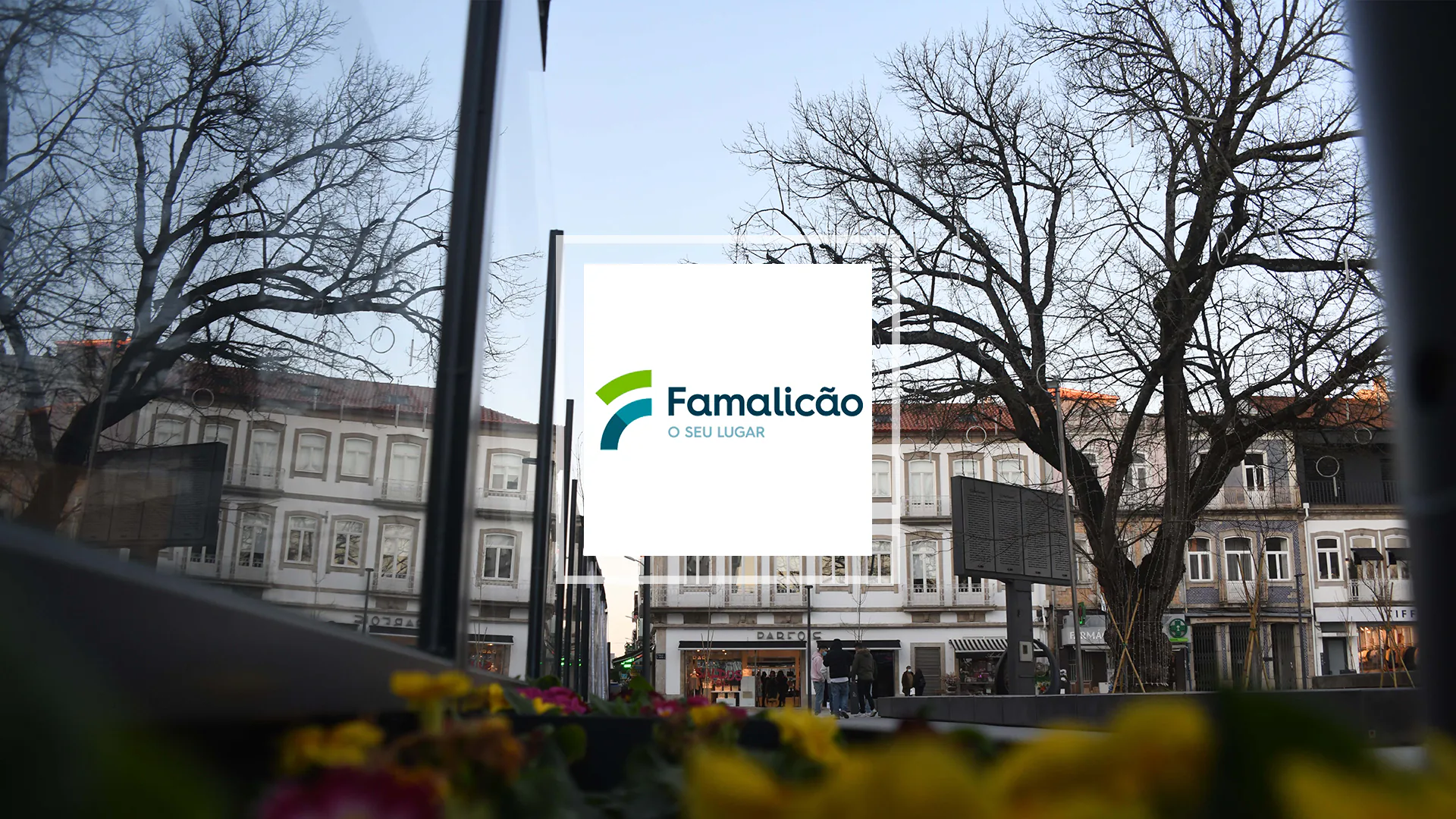 Famalicão city logo displayed over a view of a town square with historic buildings, leafless trees, and flowers in the foreground.